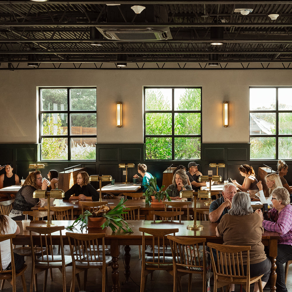 A large indoor study or library space with rows of wooden tables and chairs, many occupied by people working at desk lamps. Tall windows with dark frames line the back wall, and greenery is visible outside.