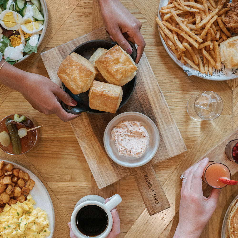 Overhead dining table scene with hands serving golden-brown biscuits from a black bowl on a wooden board, alongside fries, a side of powdered seasoning, coffee, juice, and other breakfast items.