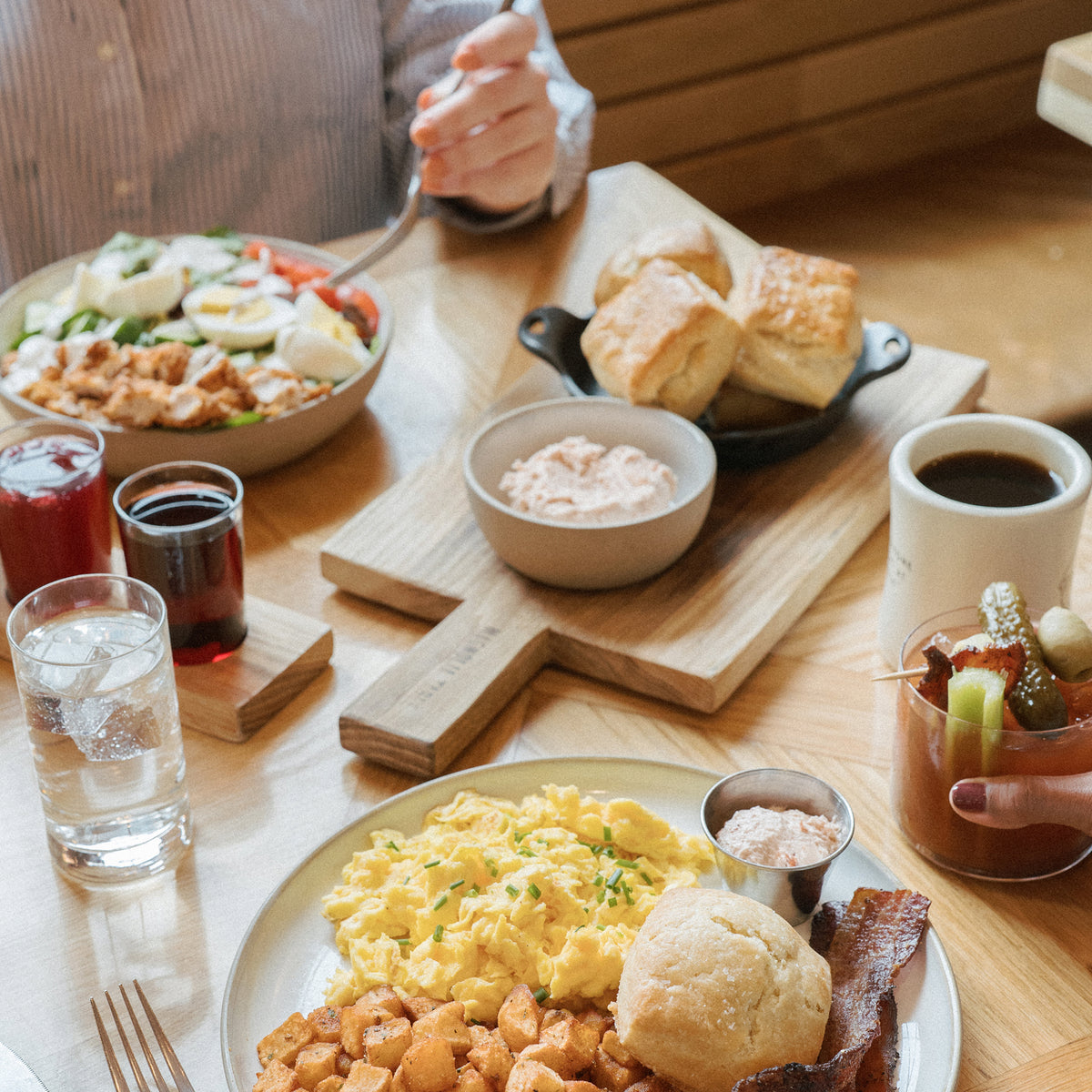 A wooden dining table set with multiple breakfast dishes, including scrambled eggs, bacon, biscuits, a salad, and coffee, with hands visible holding utensils and glasses.
