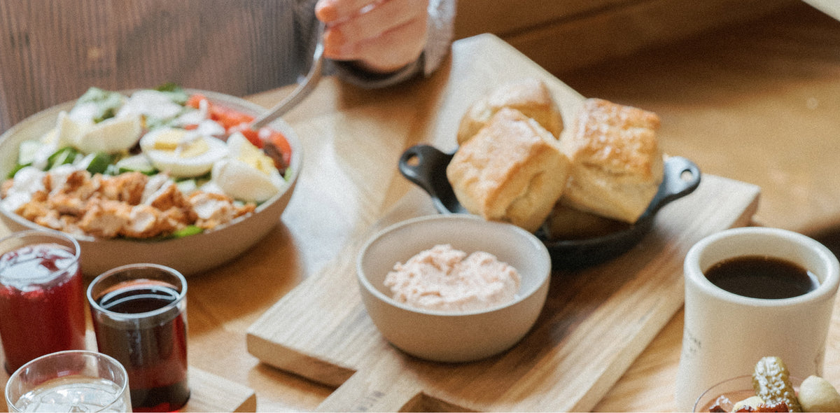 Overhead dining table scene with hands serving golden-brown biscuits from a black bowl on a wooden board, alongside fries, a side of powdered seasoning, coffee, juice, and other breakfast items.