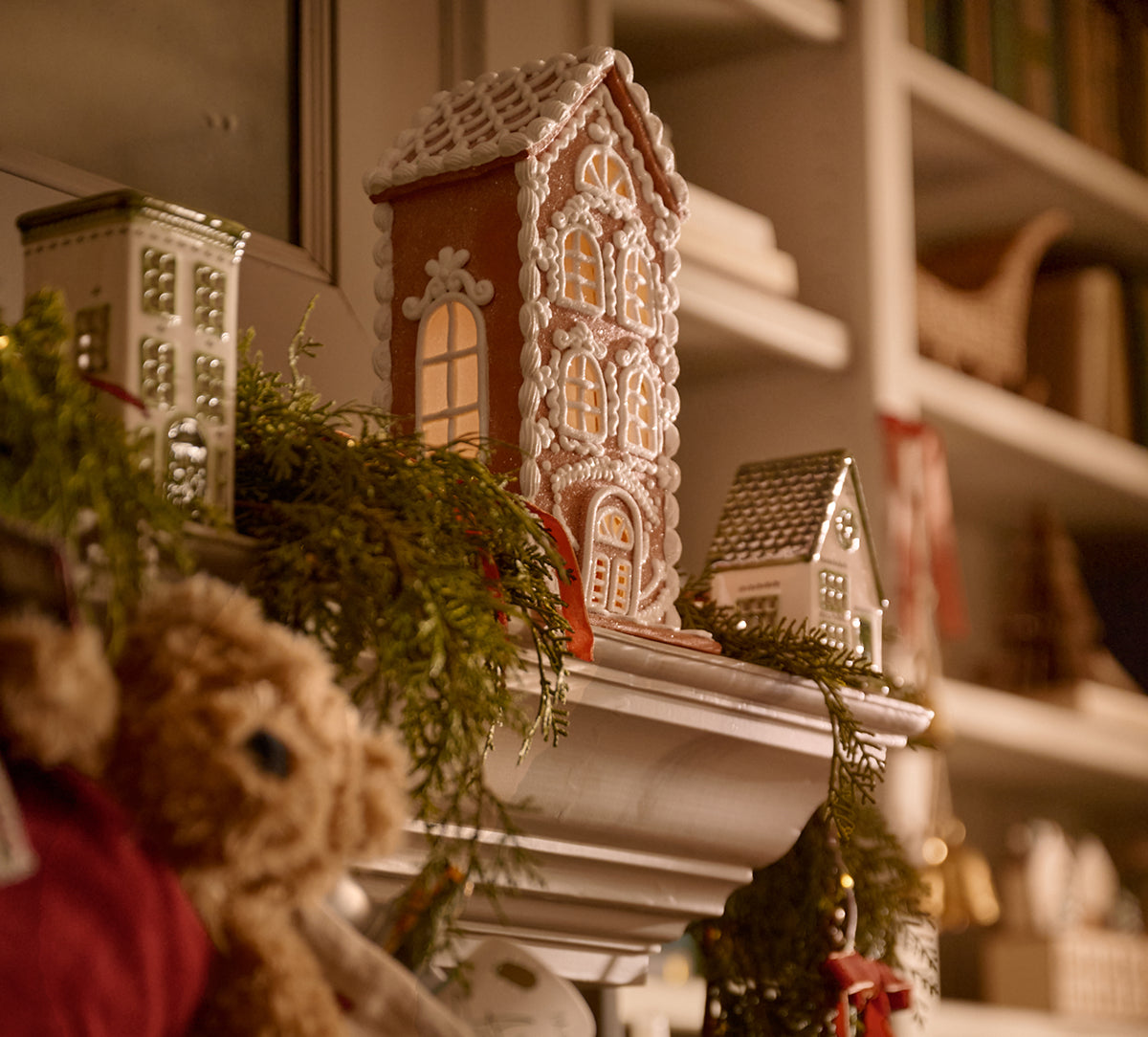 A decorated gingerbread house ornament displayed on a shelf alongside green garland, a teddy bear, and other holiday decorations in a warm indoor setting.