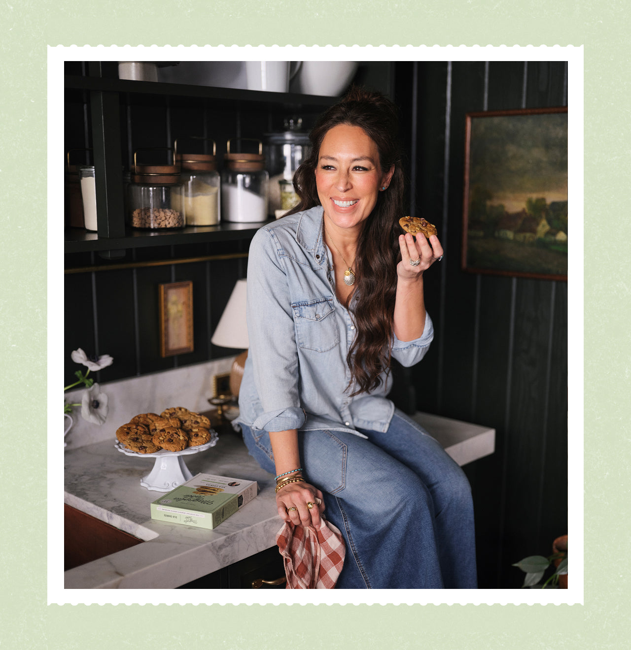 Joanna Gaines sitting on the counter in her butler's pantry holding a Magnolia Table Silos cookie in hand.  To the left is a stand full of collies and a Magnolia Table Silos Cookies box.