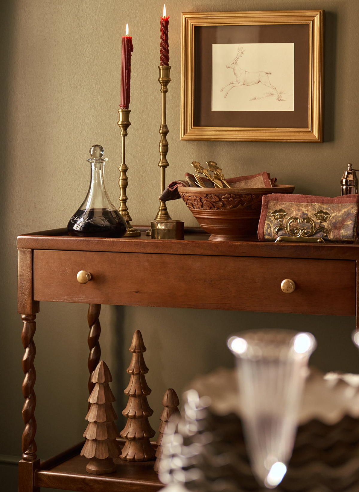 A warm, styled interior scene featuring a wooden console table with a drawer, decorated with brass candlesticks, a dark glass decanter, a carved wooden bowl, and decorative accents. A gold-framed picture hangs on the olive-green wall above.