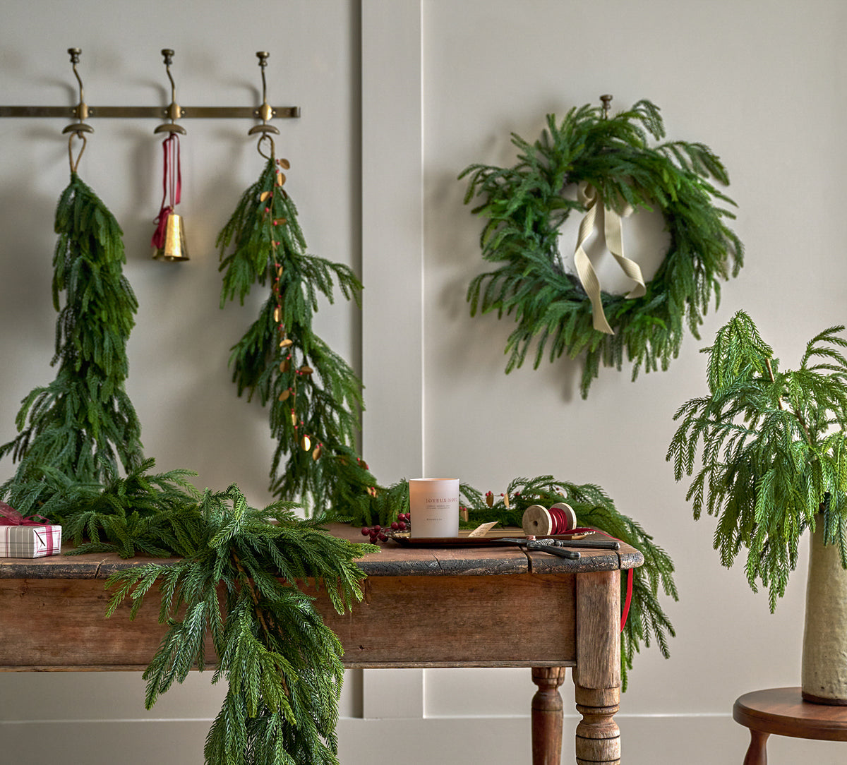 Green pine garlands and wreaths displayed in a styled interior setting, hanging on a wall and draped over a rustic wooden table with holiday decor.