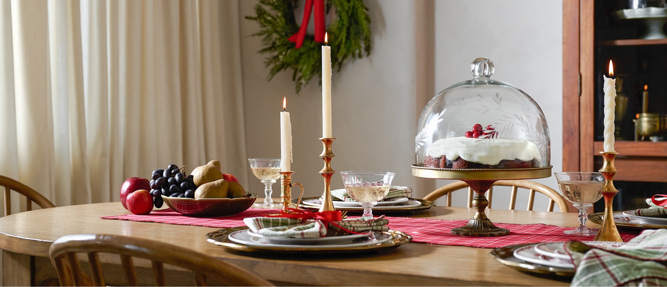 Dining table set for a festive meal with a cake under a glass cloche, candles, and fruit.