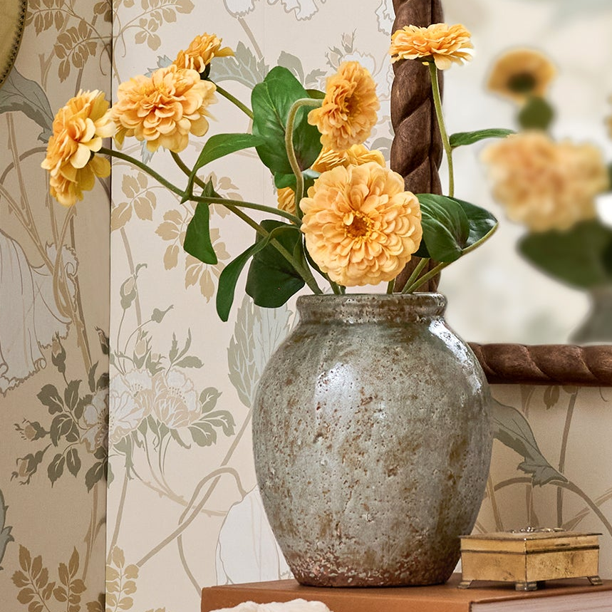 Decorative vase with yellow flowers on a wooden surface against a floral wallpaper background