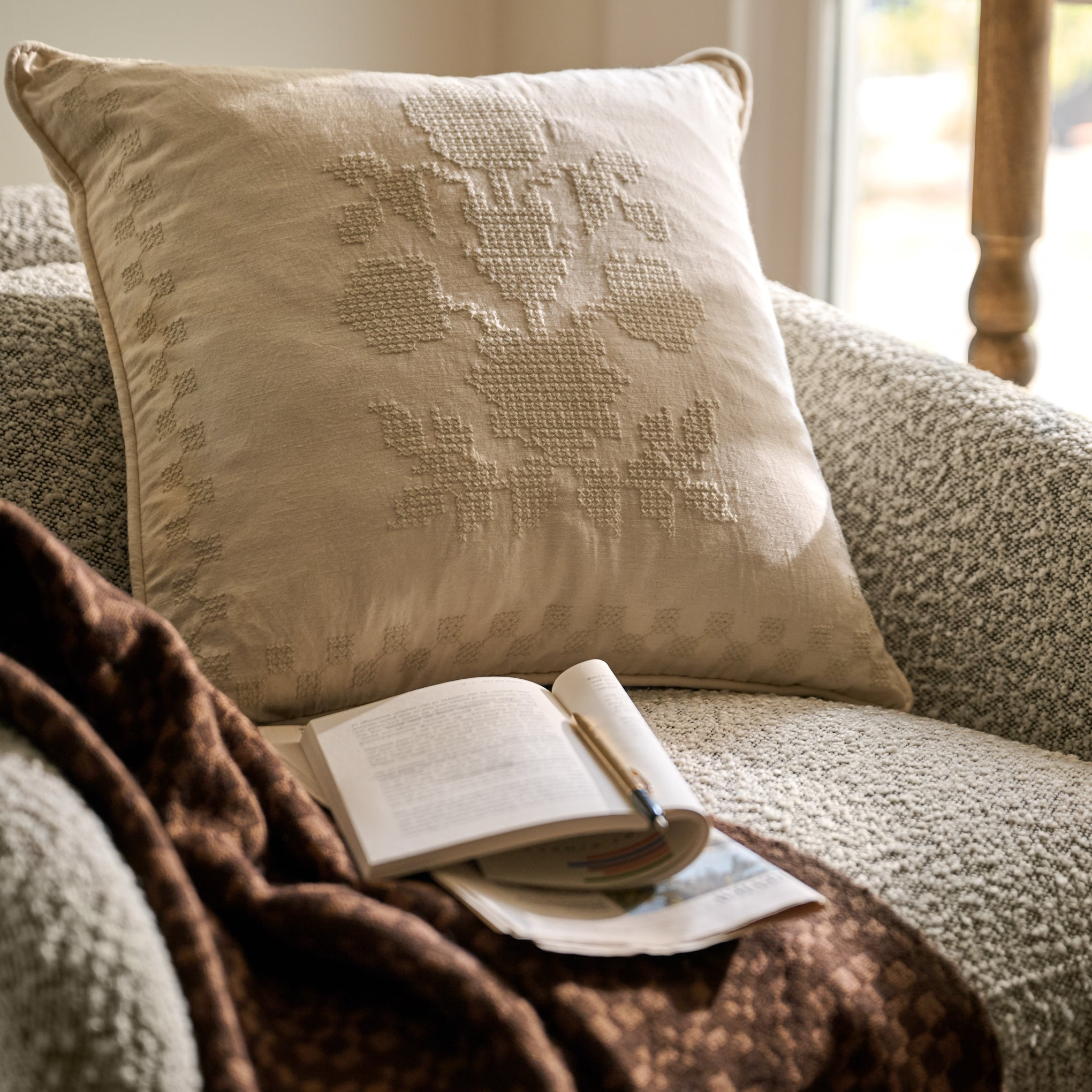 A square beige throw pillow with a textured floral cross-stitch pattern, styled on a gray armchair with a brown knit blanket and an open book.