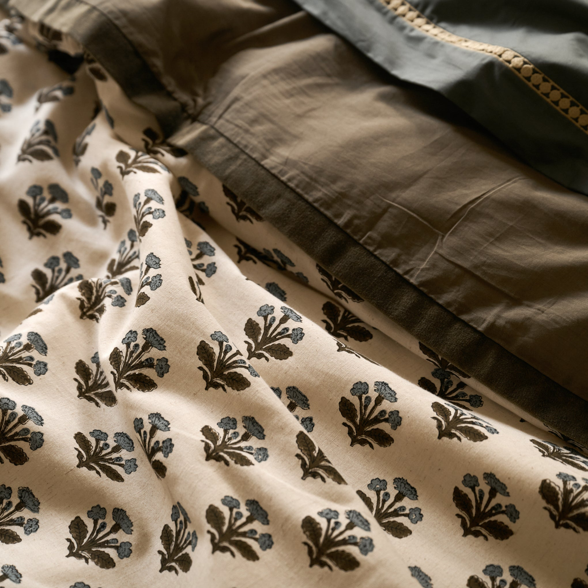 A close-up of a beige fabric with a repeating dark brown floral block print, draped alongside a solid dark olive green fabric with a gold embroidered border.