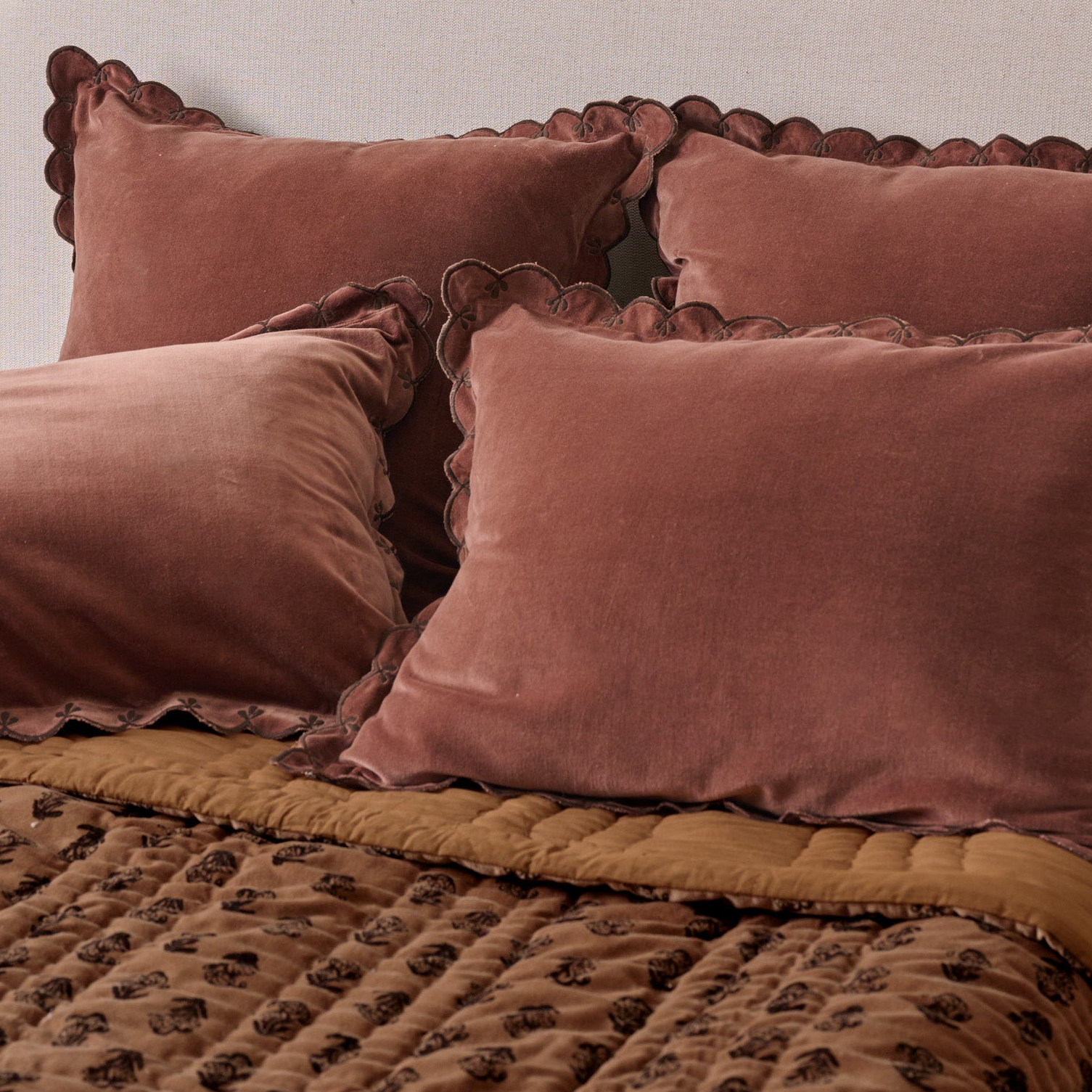 A close-up of a bed styled with dusty rose velvet pillow shams featuring scalloped edges, layered over a brown patterned quilt and a mustard yellow quilted comforter.