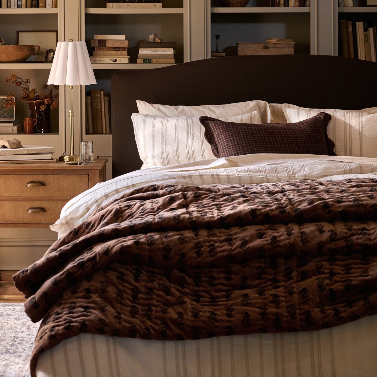 A cozy bedroom scene featuring a bed dressed in brown and cream bedding, with a patterned brown throw blanket, striped pillows, and a dark headboard. A wooden nightstand with a white lamp sits beside the bed, and bookshelves fill the background.