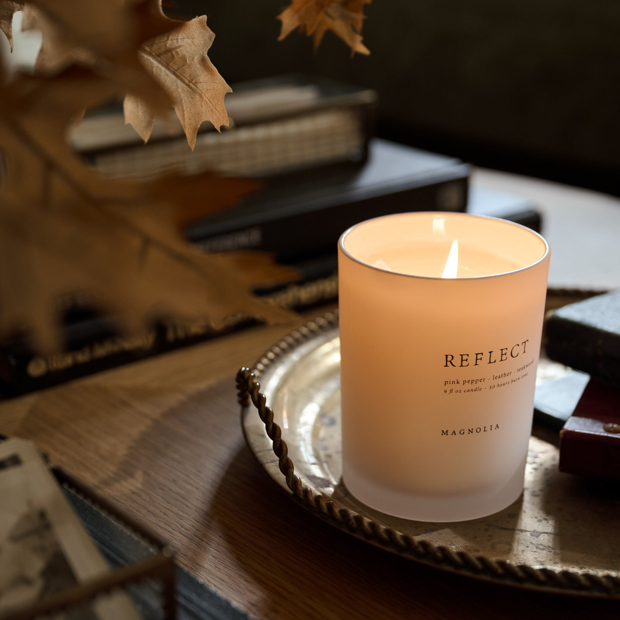 A lit scented candle in a frosted glass jar sits on a decorative tray on a wooden table, with books and dried leaves in the softly blurred background. The candle label reads 'REFLECT' and 'MAGNOLIA'.