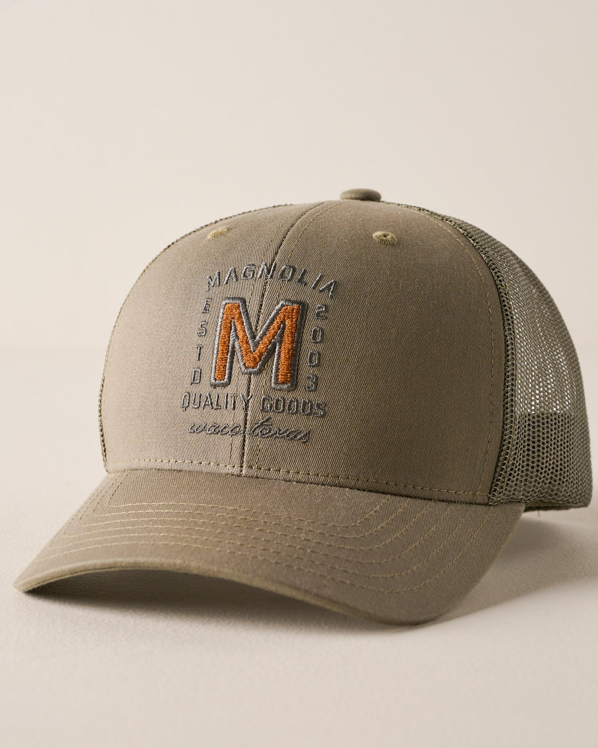 A khaki-colored trucker hat with a curved brim, featuring a mesh back and an embroidered front logo that reads 'MAGNOLIA EST. 2003 QUALITY COOLS since 2003' with a large orange 'M'.