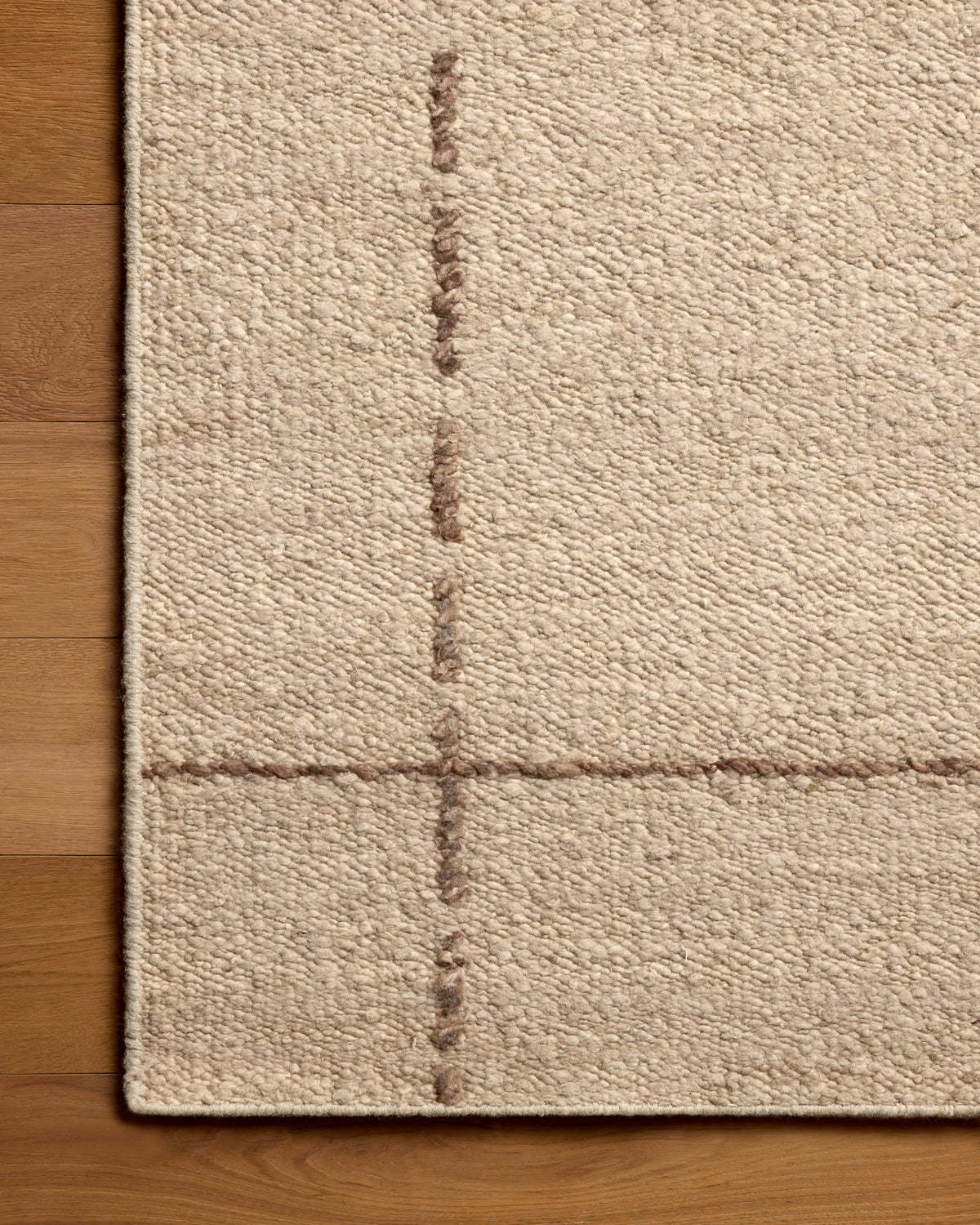 Close-up of a beige woven rug corner with a subtle stitched grid pattern, shown on a warm wood floor.