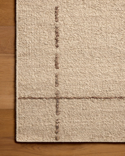 Close-up of a beige woven rug corner with a subtle stitched grid pattern, shown on a warm wood floor.