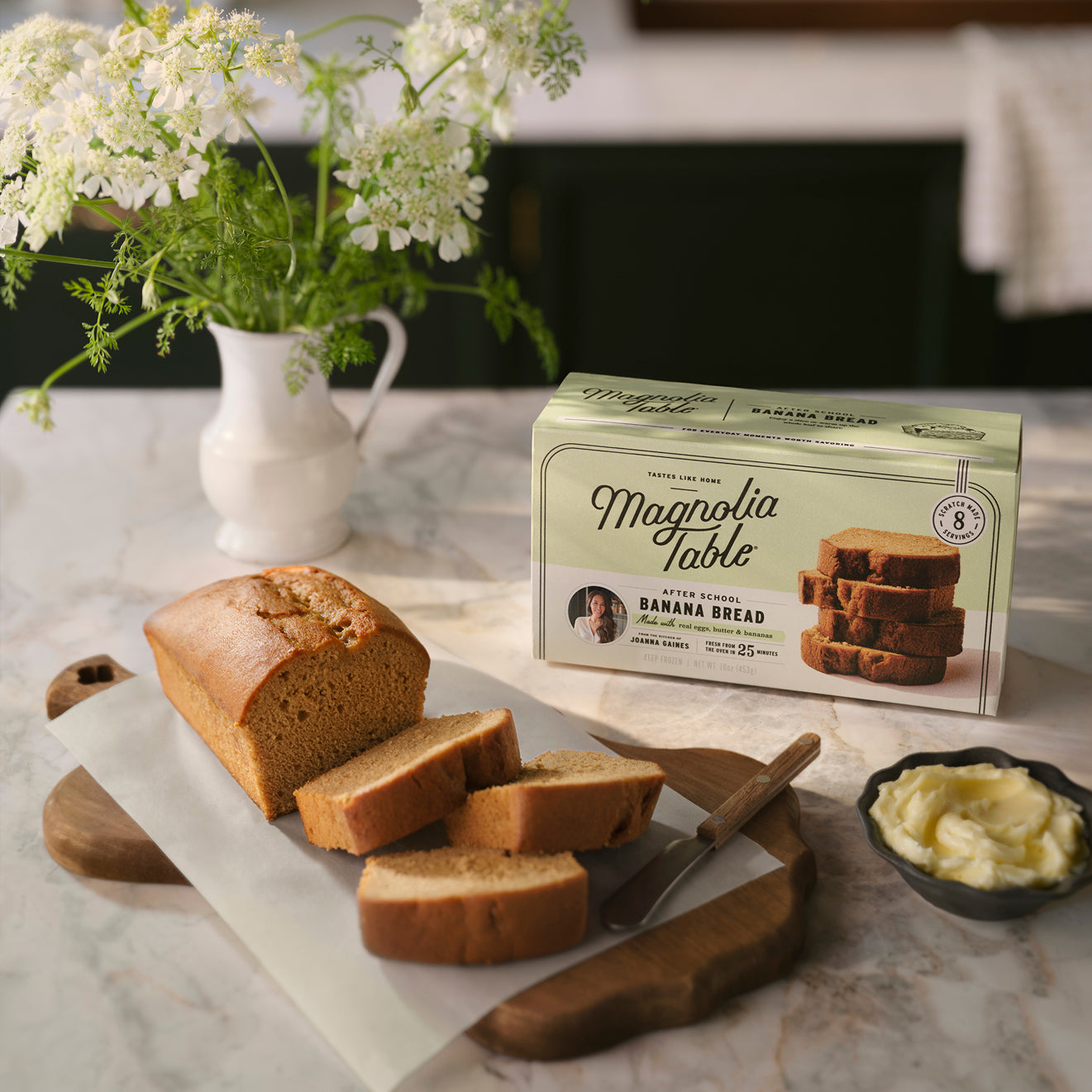 A sliced loaf of Magnolia Table After School Banana Bread on a cutting board with a box of Magnolia Table Frozen After School Banana Bread and flowers in the background.