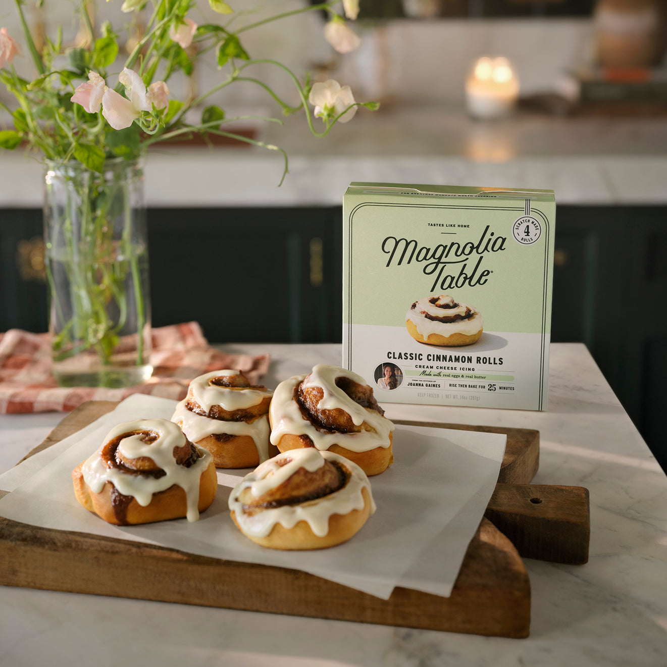 Magnolia Table Classic Cinnamon Rolls with cream cheese icing on a wooden board next to a box of Magnolia Table Frozen Classic Cinnamon Rolls. 