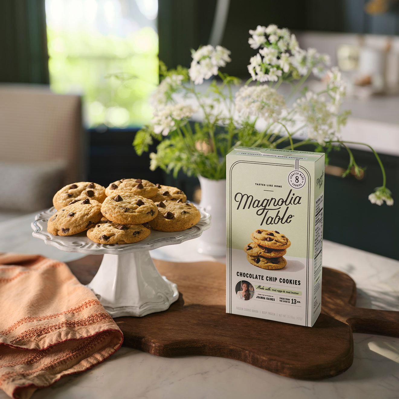 Box of Magnolia Table Frozen Chocolate Chip Cookie Dough on a kitchen island with Magnolia Table Chocolate Chip Cookies on a plate stand and flowers in the background.