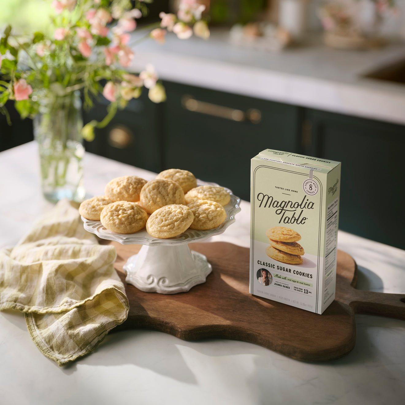 Box of Magnolia Table Frozen Classic Sugar Cookie Dough on a kitchen island with Magnolia Table Classic Sugar Cookies on a plate stand and flowers in the background.