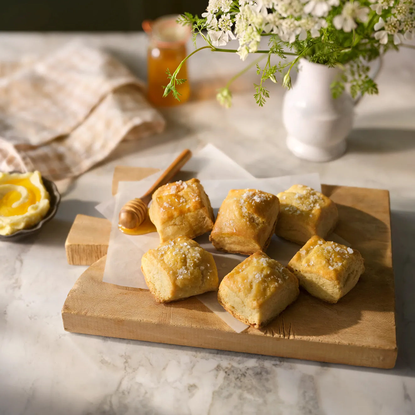Wooden cutting board with Magnolia Table Jo's Buttermilk Biscuits, a honey dipper, and a vase of flowers on a marble surface.