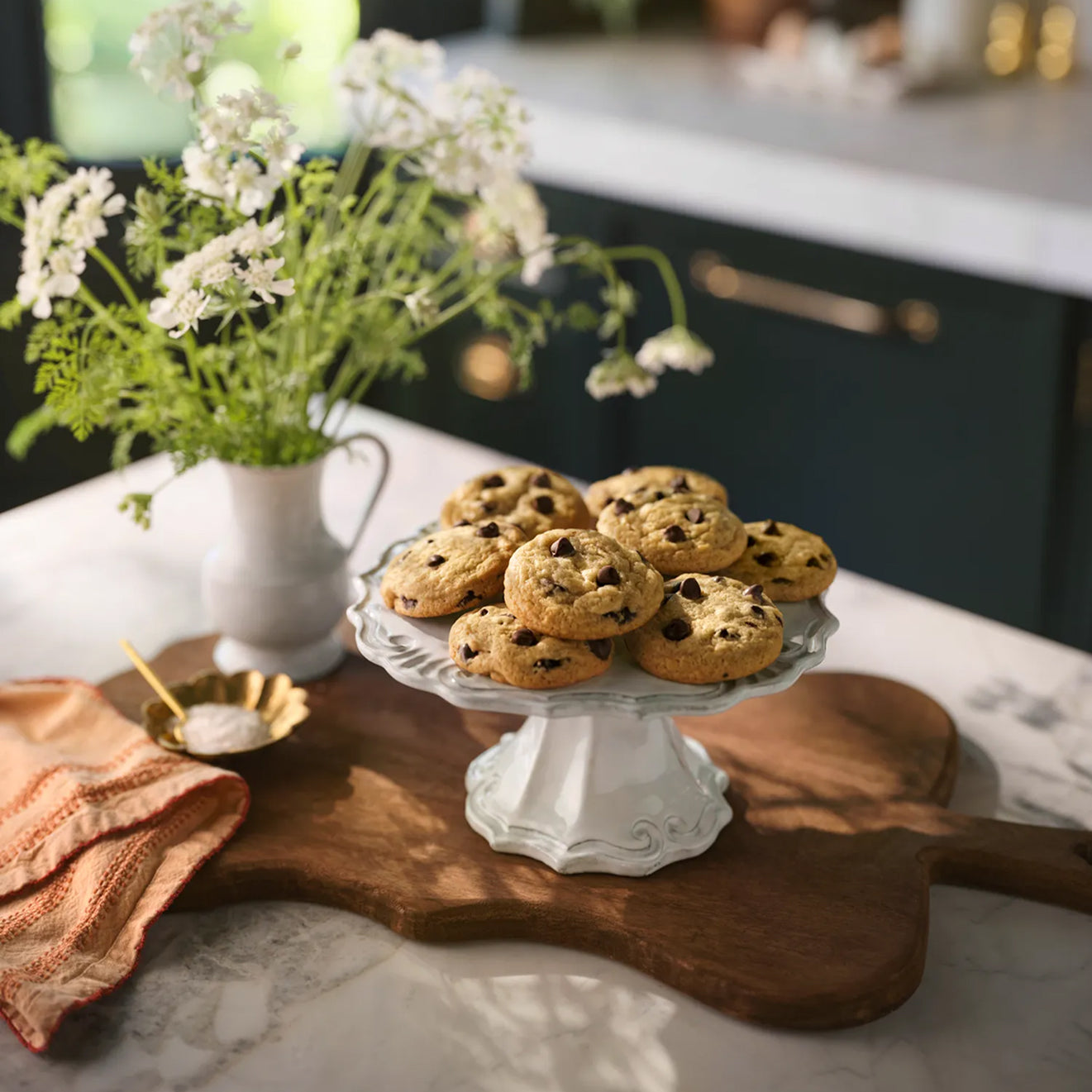 Magnolia Table Chocolate Chip Cookies on a white cake stand with a vase of flowers and wooden cutting board in the background.
