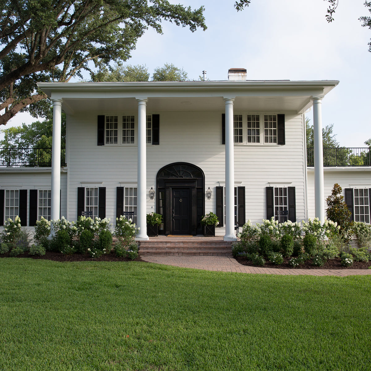 A classic white colonial-style house with black shutters, a front porch supported by tall white columns, a green lawn, and landscaped flower beds in the foreground.