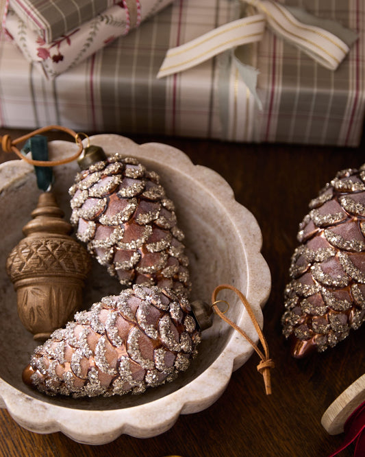 Decorative pinecones with glitter and an acorn in a white dish, with a gift box in the background.
