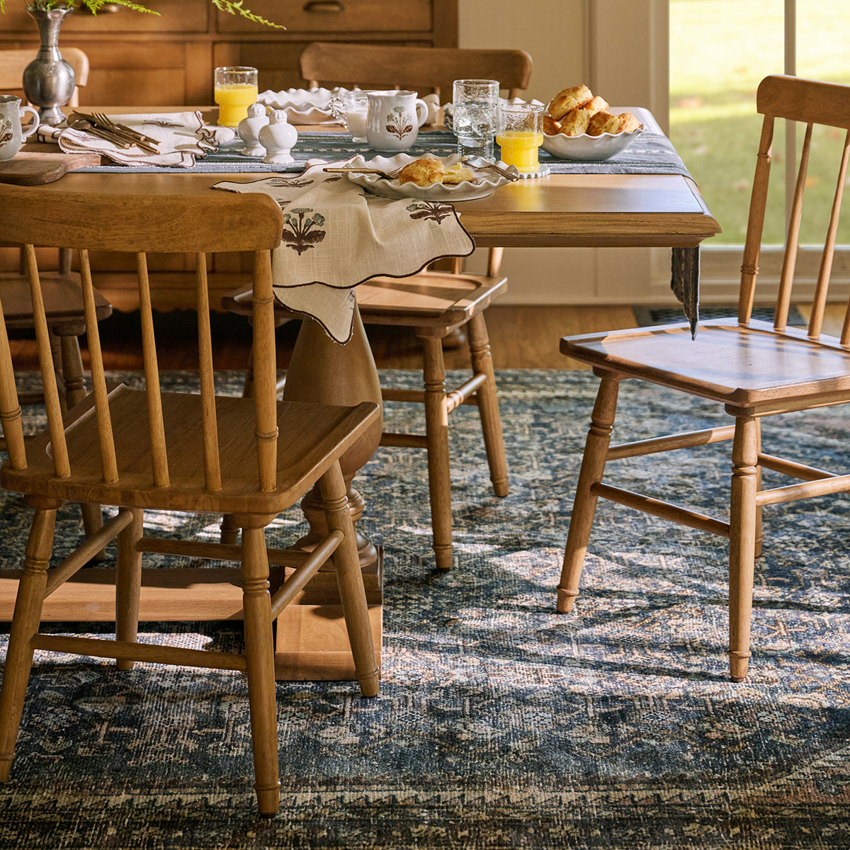 A styled dining room scene with a wooden dining table and spindle-back chairs on a blue patterned area rug, set with breakfast tableware and food near a window.