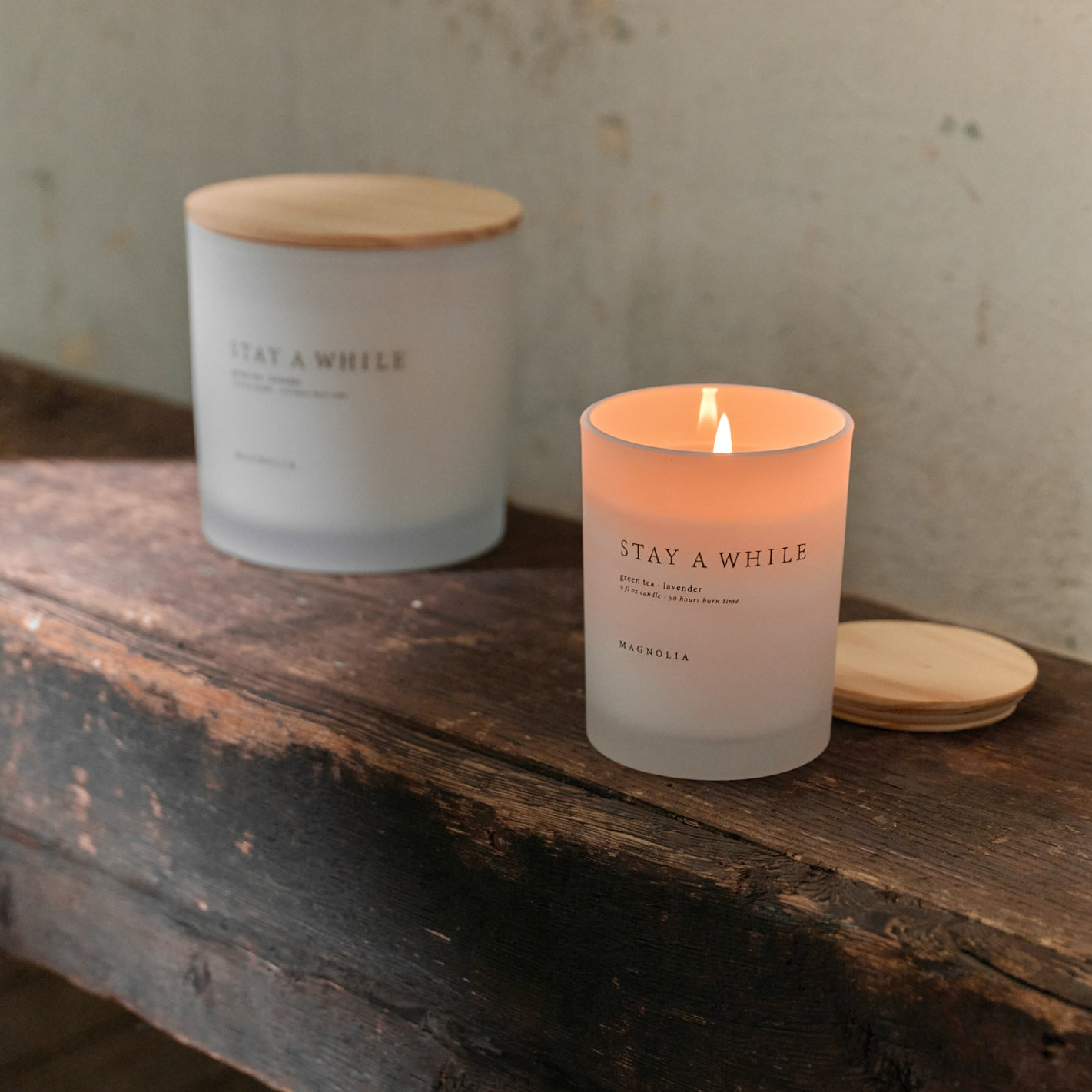 Two magnolia signature candles sitting on top an old wooden bench. The larger size of the "Stay a While" glass candle is in the background, with the smaller version in front lit for ambiance. 