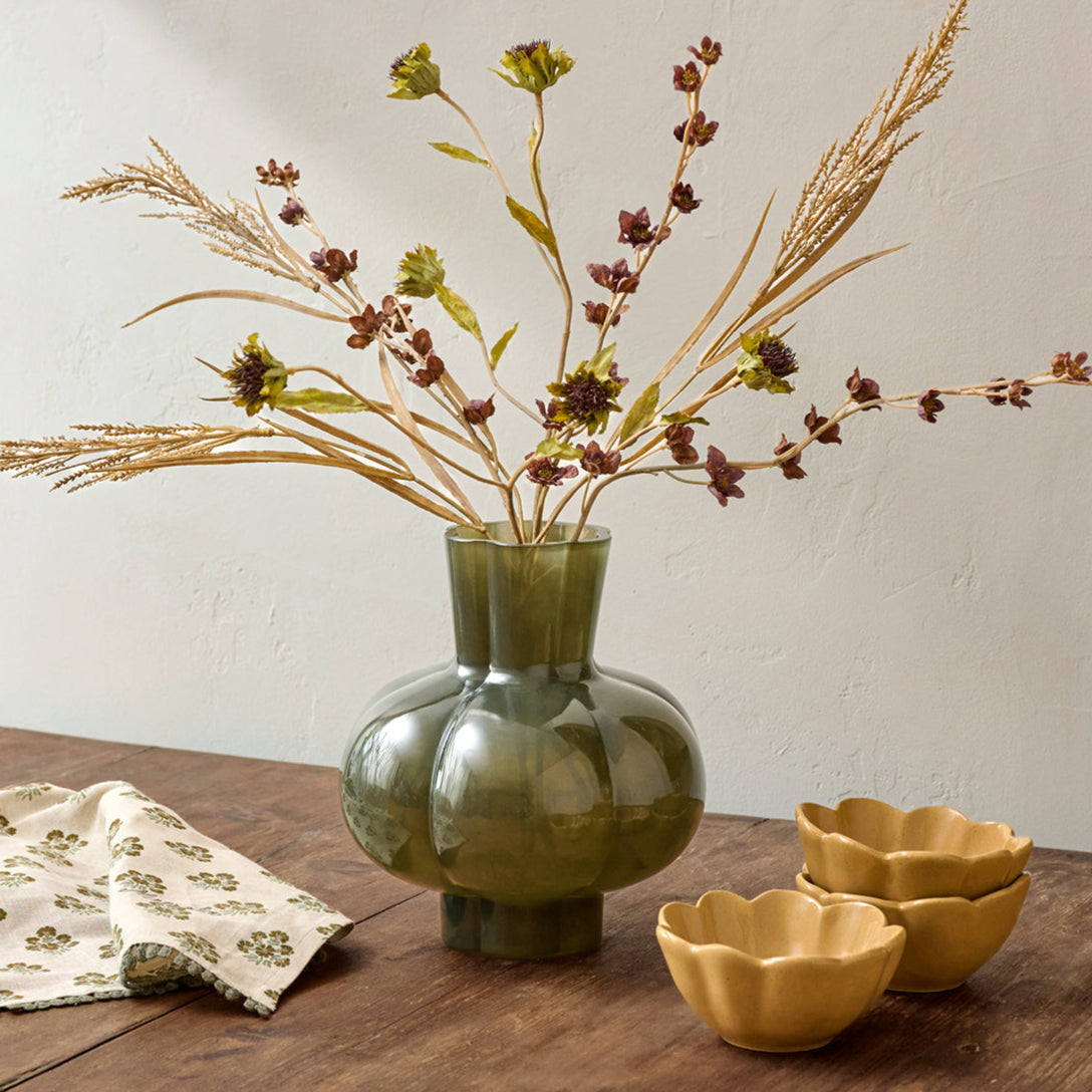 Green vase with dried flowers on a wooden table with yellow bowls and a patterned cloth.