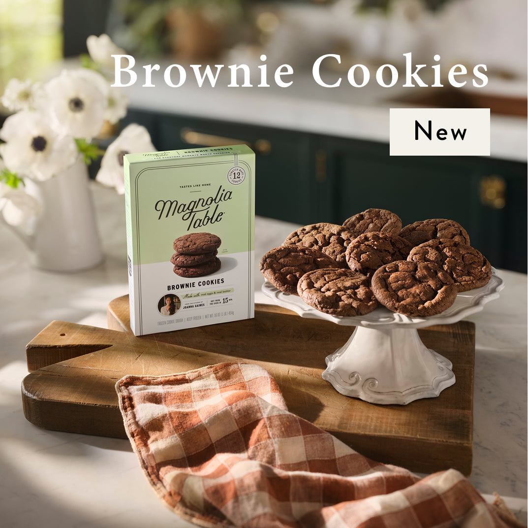 Magnolia Table Brownie cookies on a white stand with a box labeled 'Magnolia Table Brownie Cookies' on a wooden cutting board.