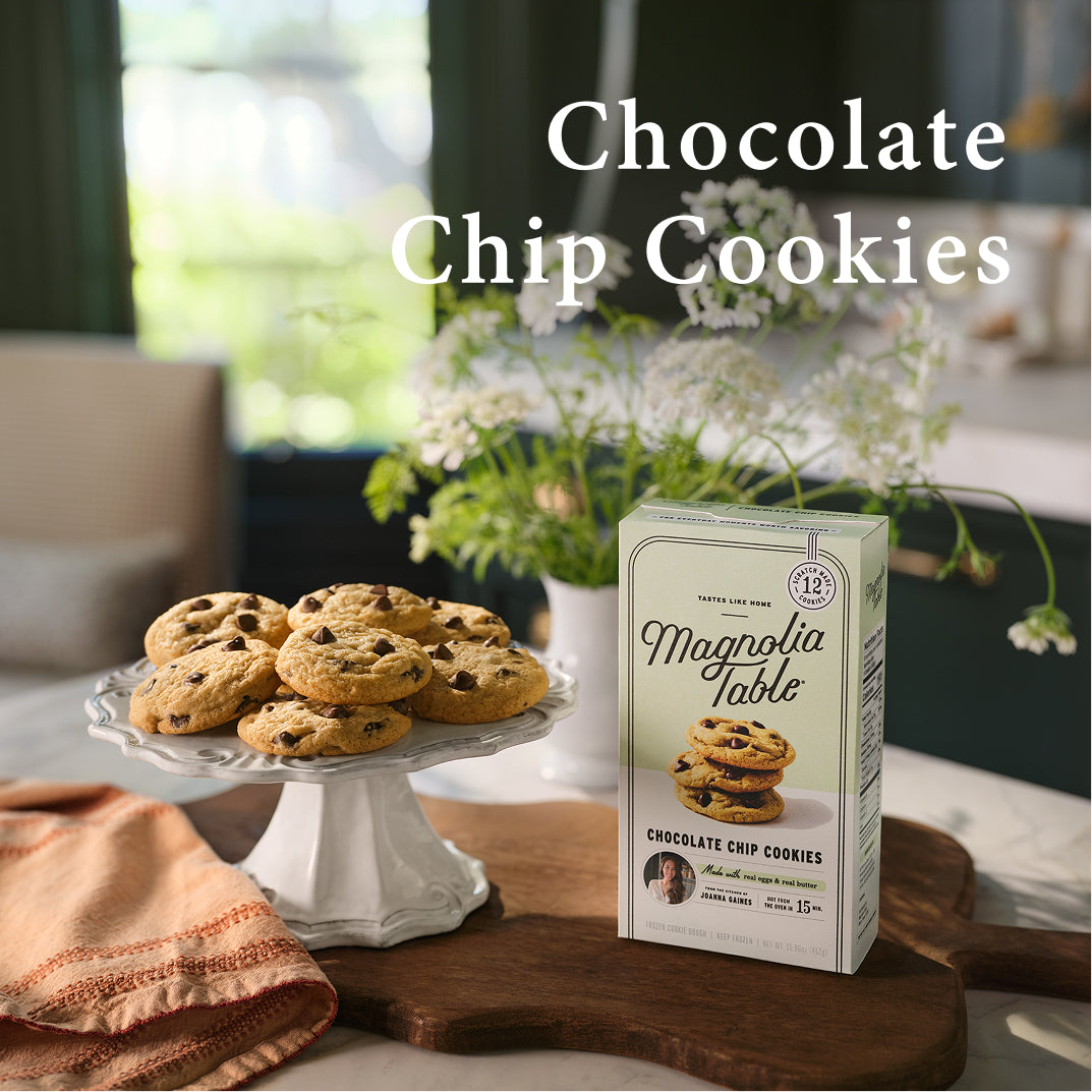 Box of Magnolia Table chocolate chip cookies on a table with chocolate chip cookies on a stand.