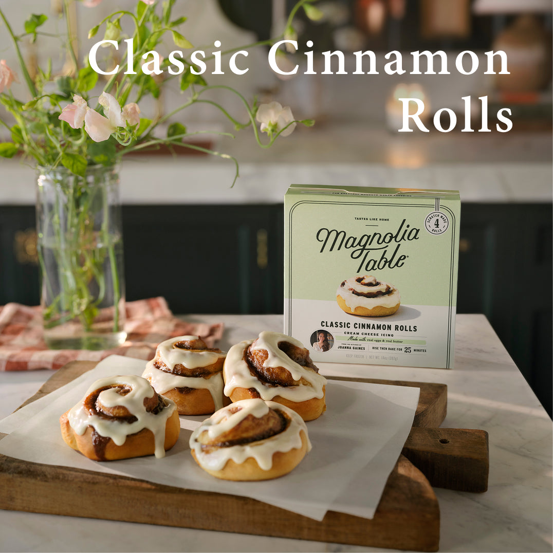 Magnolia Table Cinnamon rolls on a wooden board with a box of Magnolia Table cinnamon rolls in the background.