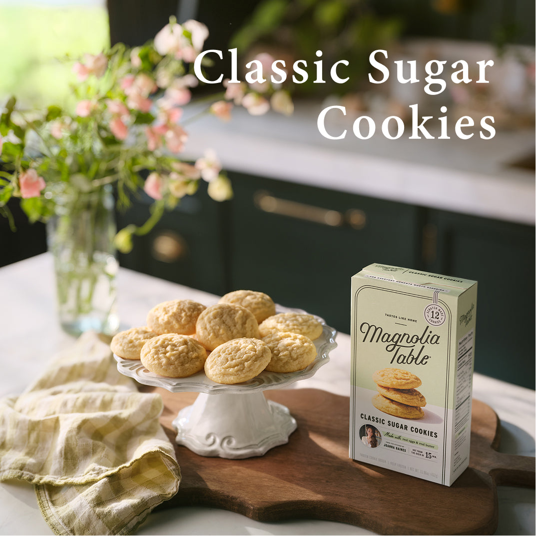 Box of Magnolia Table Classic Sugar Cookies on a kitchen counter with classic sugar cookies and flowers.
