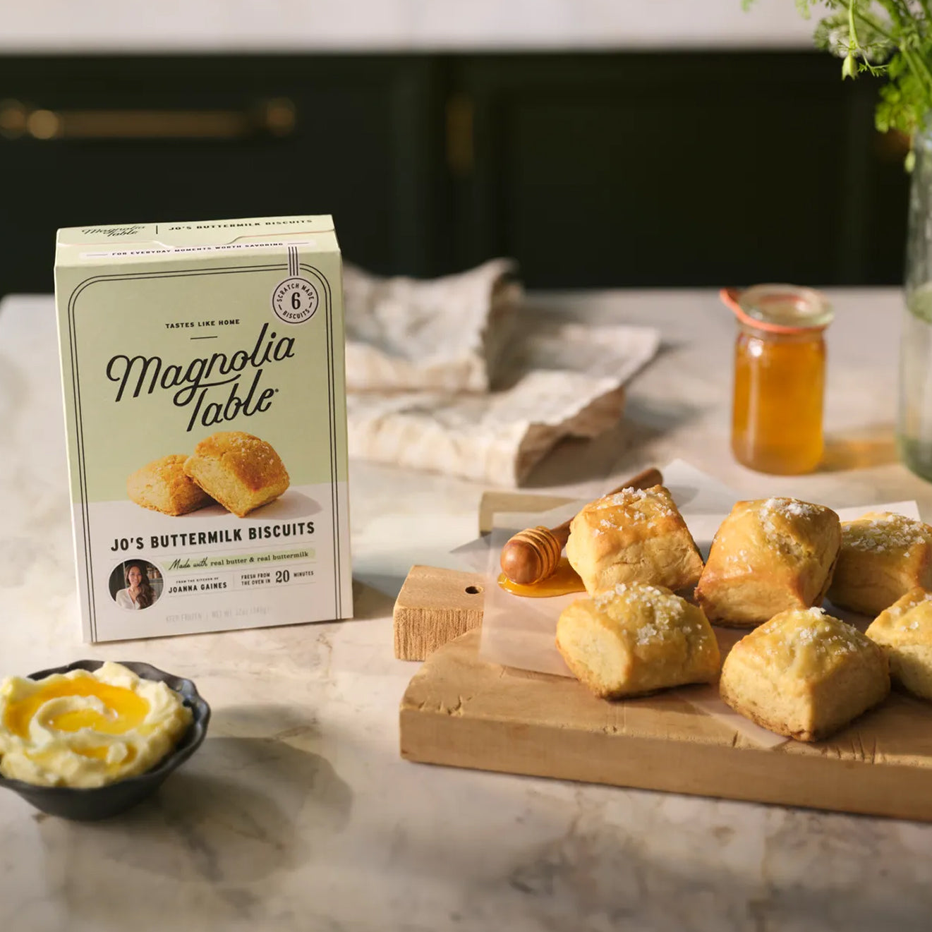 Box of Magnolia Table Frozen Jo's Buttermilk Biscuits on a marble countertop with Magnolia Table Jo's Buttermilk Biscuits and honey on a wooden cutting board.