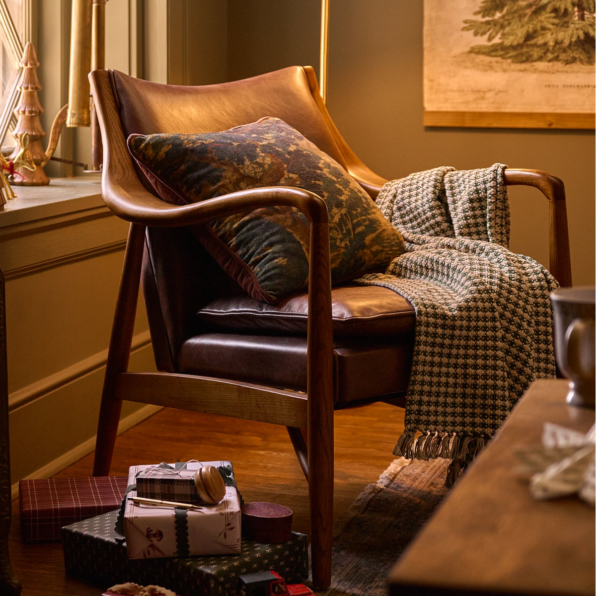 A mid-century modern leather armchair in a warm, cozy room setting, styled with a patterned throw pillow, a checkered knit blanket, and holiday gift boxes on the floor.