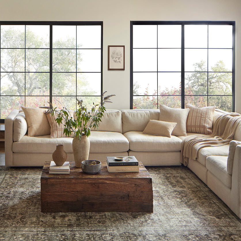 A large cream sectional sofa in a bright, modern living room with black-framed windows, a rustic wooden coffee table, and neutral-toned decor.