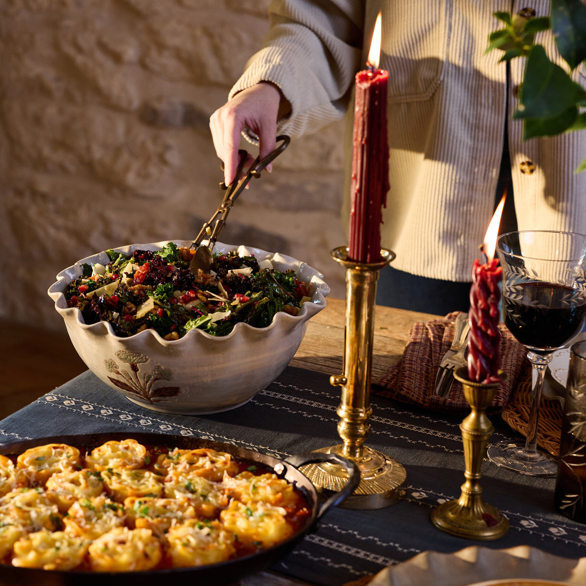 A person serving food from a large ceramic bowl on a rustic table set with lit red candles, a glass of red wine, and a pan of stuffed pasta. The person is wearing a cream ribbed sweater.