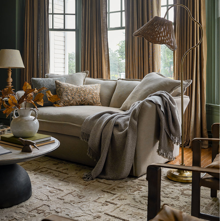 A beige upholstered sofa in a styled living room, adorned with neutral throw pillows, a cream knit throw blanket, and a wicker floor lamp, set against dark green curtains and wood flooring.