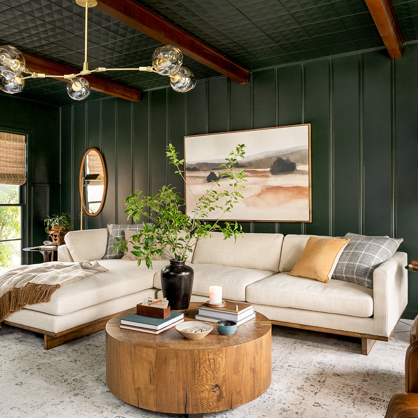 A modern living room featuring a large cream sectional sofa with a round wooden coffee table in the foreground, set against dark green paneled walls and a dark green textured ceiling with exposed wood beams.