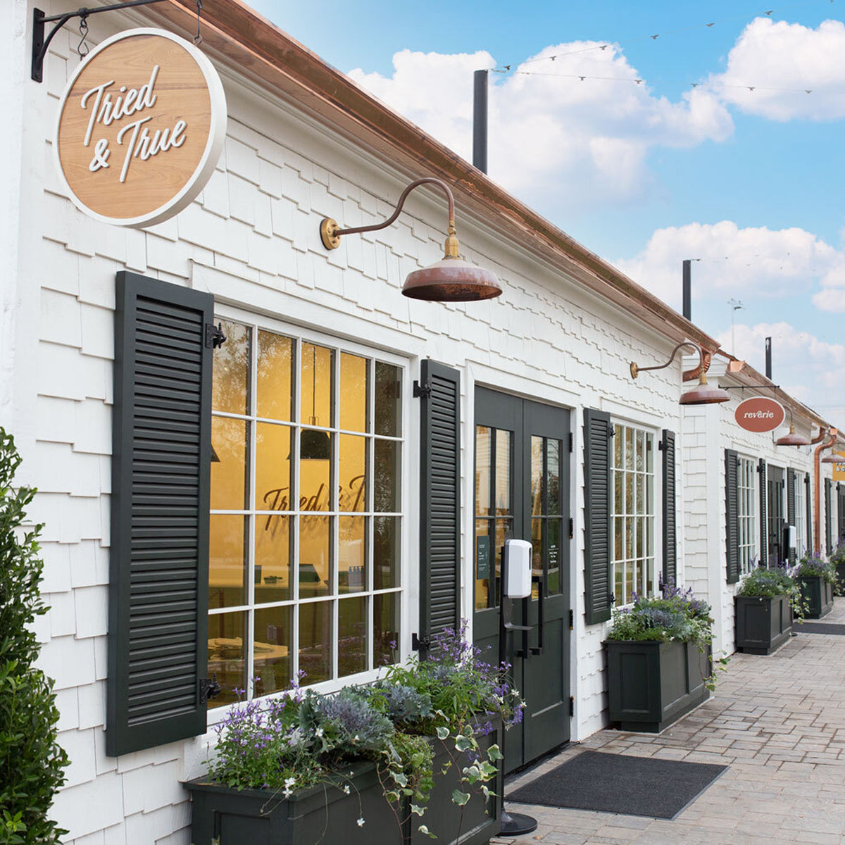 A white shingled storefront building with black shutters, a round wooden 'Tried & True' sign, outdoor wall lamps, planters, and a paved walkway under a blue sky.