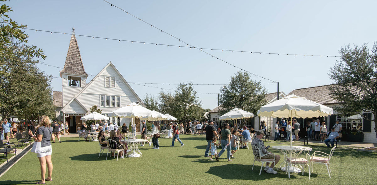 An outdoor patio scene on a grassy lawn with many people dining under white umbrellas beside a white building, with string lights overhead and trees in the background.