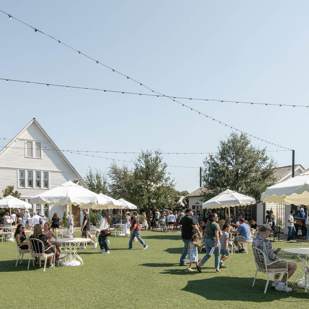 An outdoor patio scene on a grassy lawn with many people dining under white umbrellas beside a white building, with string lights overhead and trees in the background.