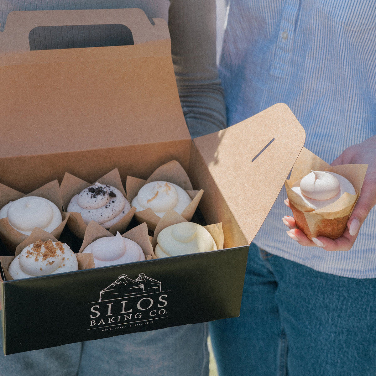 An open kraft paper bakery box from Silos Baking Co. holds several frosted cupcakes, while a person in a blue striped shirt holds one cupcake outside in natural light.