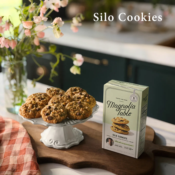 Box of Magnolia Table Frozen Silo Cookies on a kitchen island with baked silo cookies on a plate stand and flowers.  Silo Cookies.  Learn More.