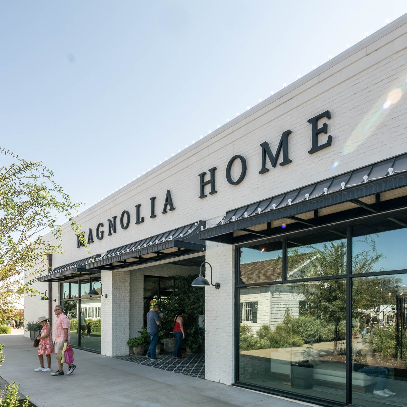 A white brick home exterior with large black lettering reading 'MAGNOLIA HOME' above a storefront with large glass windows. A few people are walking and standing at the entrance, and trees and another house are visible in the background.
