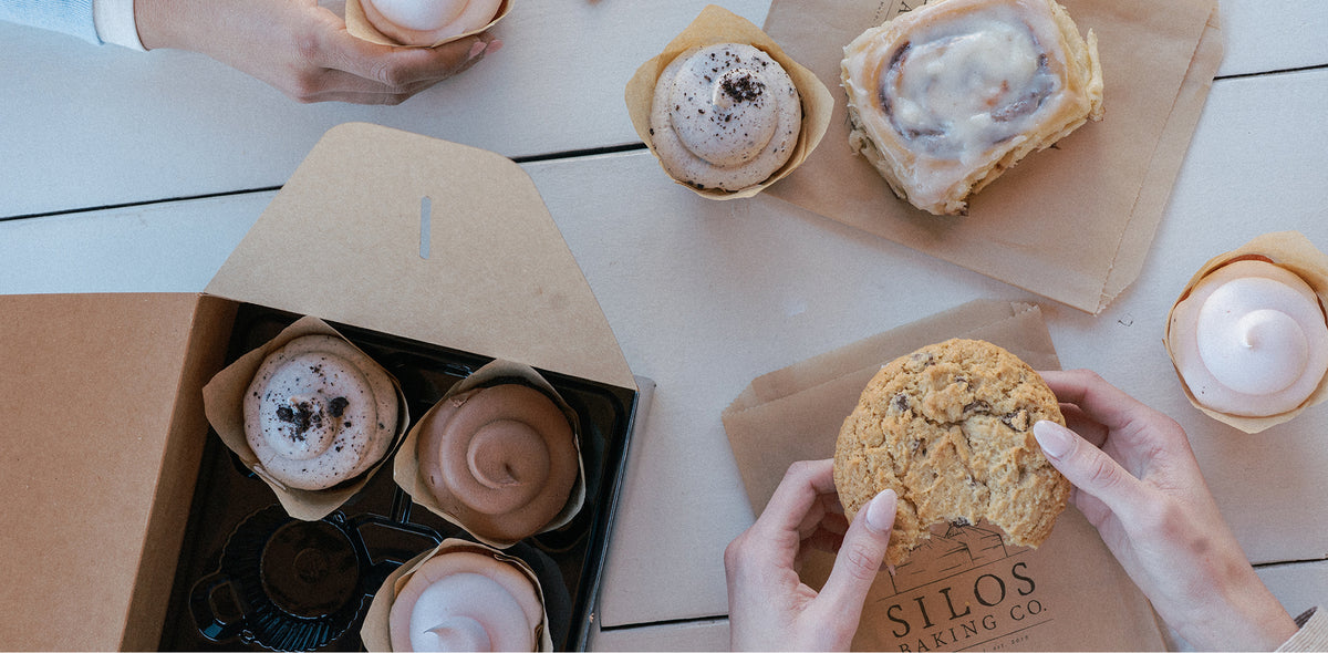 Overhead view of assorted baked goods on a light tabletop: several frosted cupcakes, a cinnamon roll, a chocolate chip cookie being held in hands, and a black bakery box. A paper bag printed with 'SILOS BAKING CO' is visible.