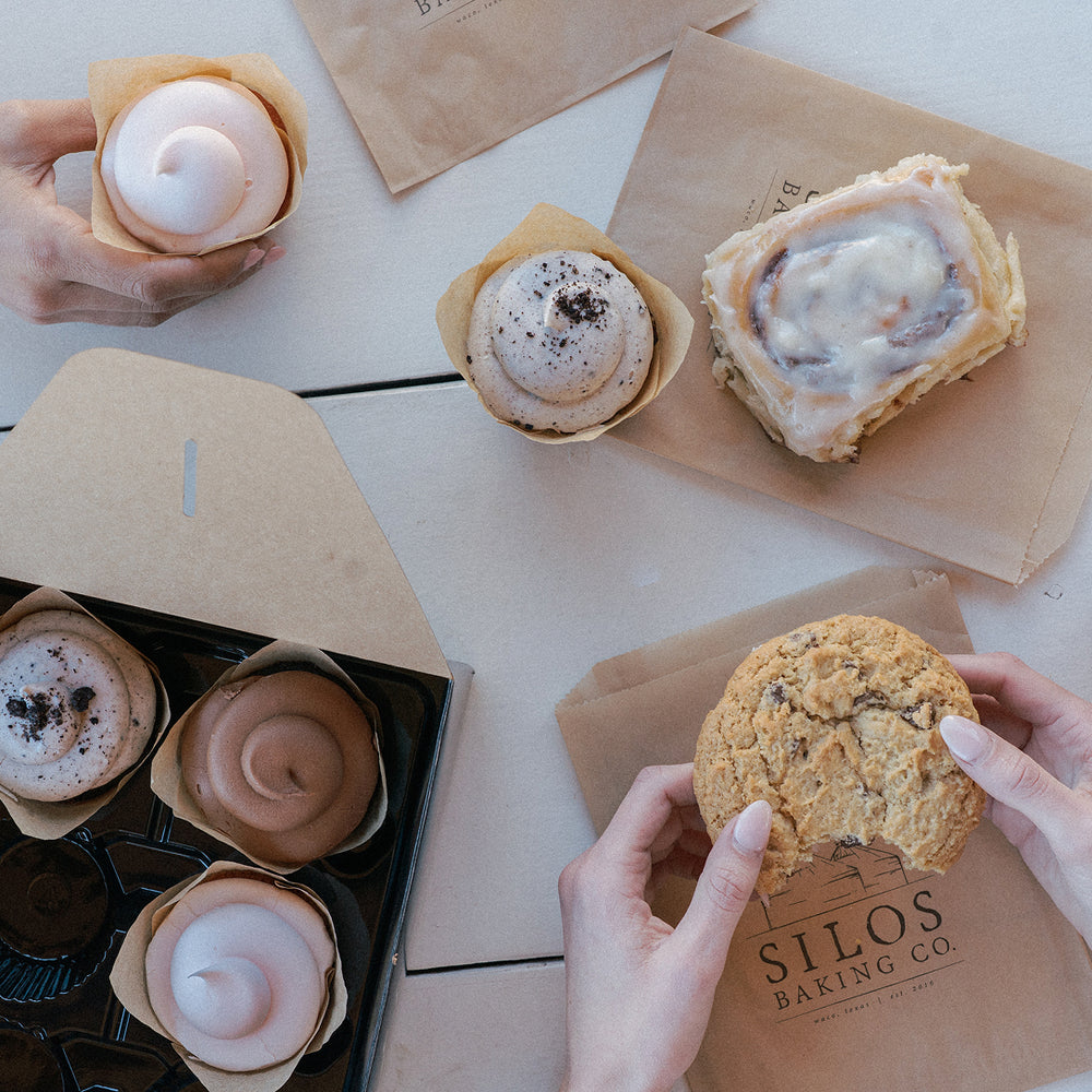 Overhead view of assorted baked goods on a light tabletop: several frosted cupcakes, a cinnamon roll, a chocolate chip cookie being held in hands, and a black bakery box. A paper bag printed with 'SILOS BAKING CO' is visible.