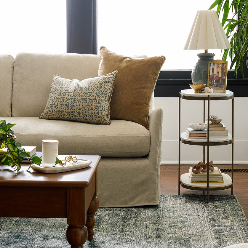 A beige slipcovered sofa in a styled living room, decorated with two throw pillows, a wooden coffee table, a two-tier side table with a lamp, and a patterned area rug.