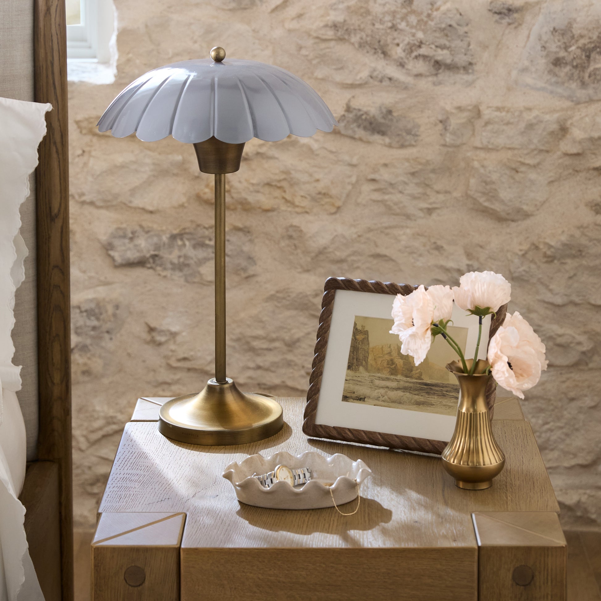 Carolyn Table lamp on a nightstand with a vase of flowers and framed picture against a stone wall.