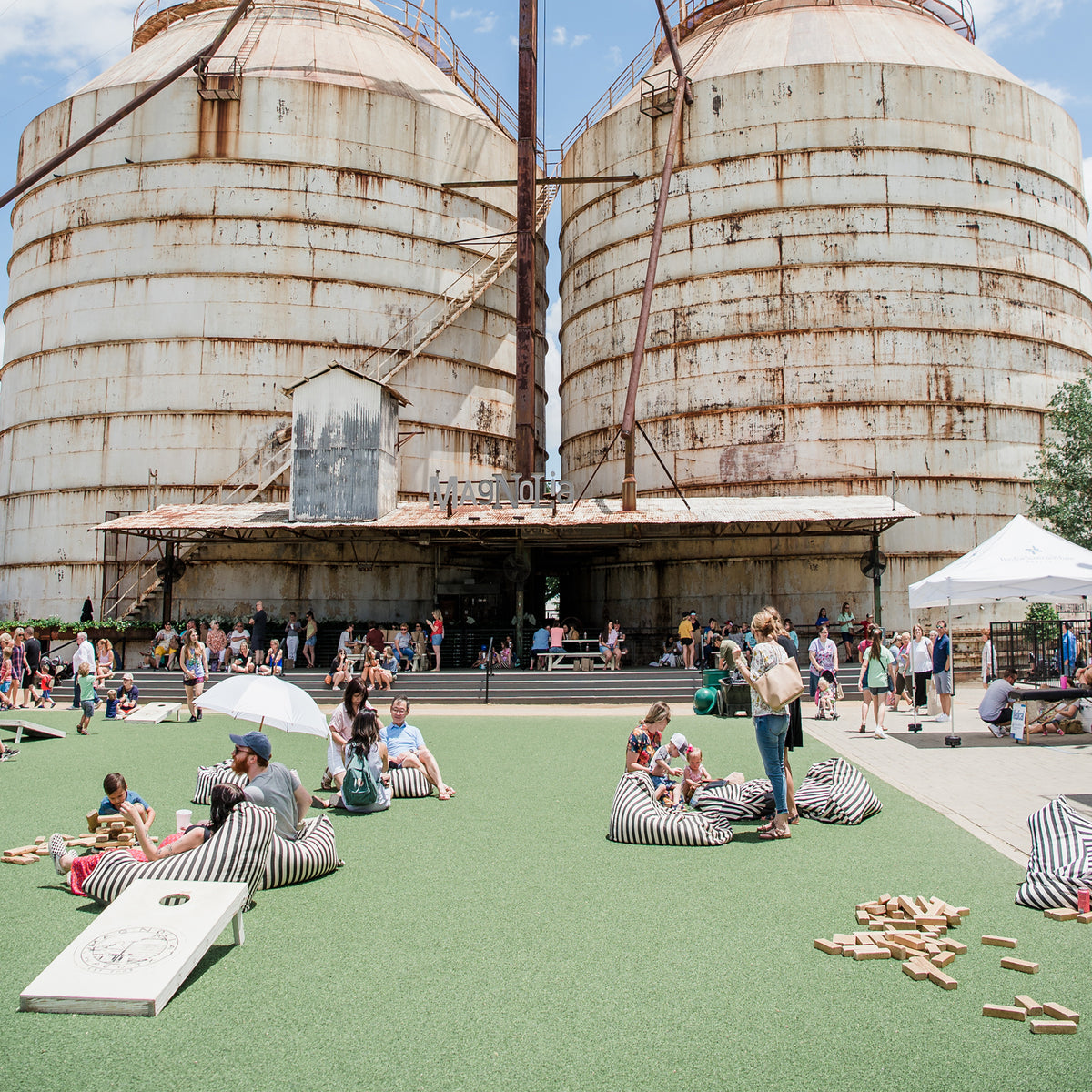 Outdoor scene in front of two large rusted industrial silos, with many people sitting on striped bean bags on green artificial turf, playing games, and walking near a covered walkway and white tent.